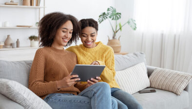 Two African American female sitting on sofa in living room surfing on a tablet